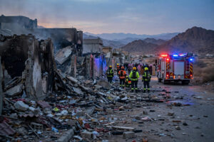 Damaged residential street in Najran, Saudi Arabia, after a Houthi projectile strike, with civil-defense crews and smoke at dusk
