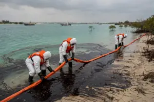 Emergency workers in protective gear deploy an orange containment boom along an oiled beach in Umm Al Quwain, UAE, as dark oil spreads across turquoise Gulf water.