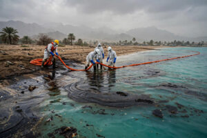 Workers in protective gear deploy an orange containment boom along an oiled beach in Ras Al Khaimah, UAE, as dark oil spreads across turquoise Gulf water
