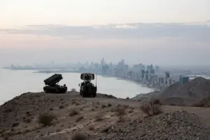 Saudi air defence battery silhouetted on a desert ridge above an Eastern Province coastal city skyline at dawn